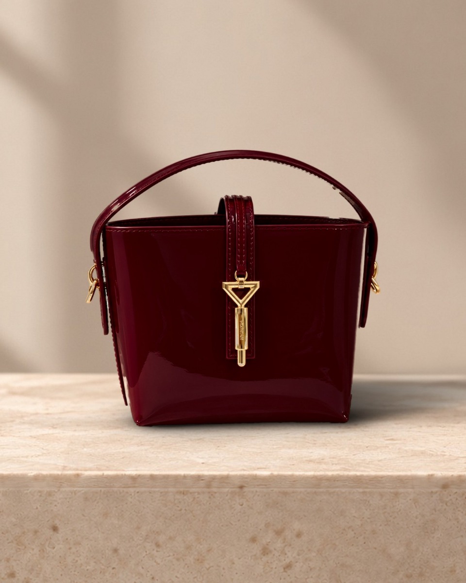 Closer view of the Alaina Mini Bucket Bag: glossy burgundy patent and gold triangle clasp engraved FLORIAN, in warm window light