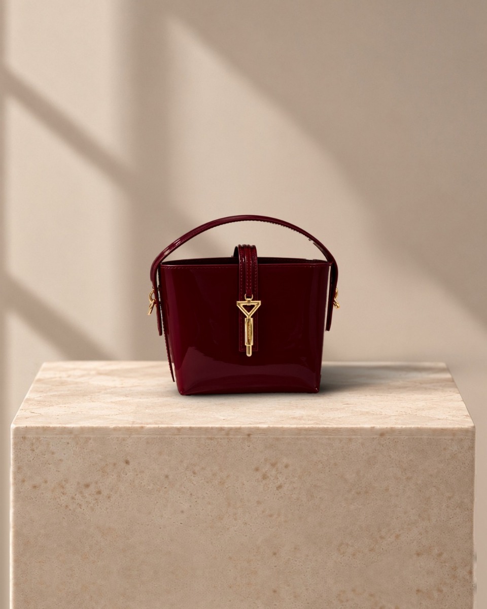 Alaina Mini Bucket Bag in deep burgundy patent leather with gold triangle clasp, on a travertine plinth in warm window light