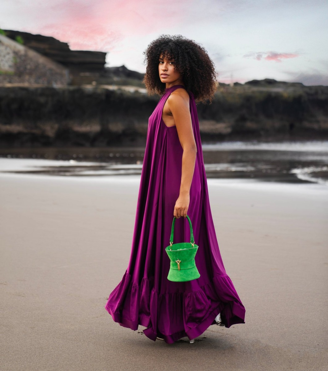A model in a flowing purple satin dress on a beach at dusk, carrying a bespoke green suede Florian bucket bag with a gold-tone clasp
