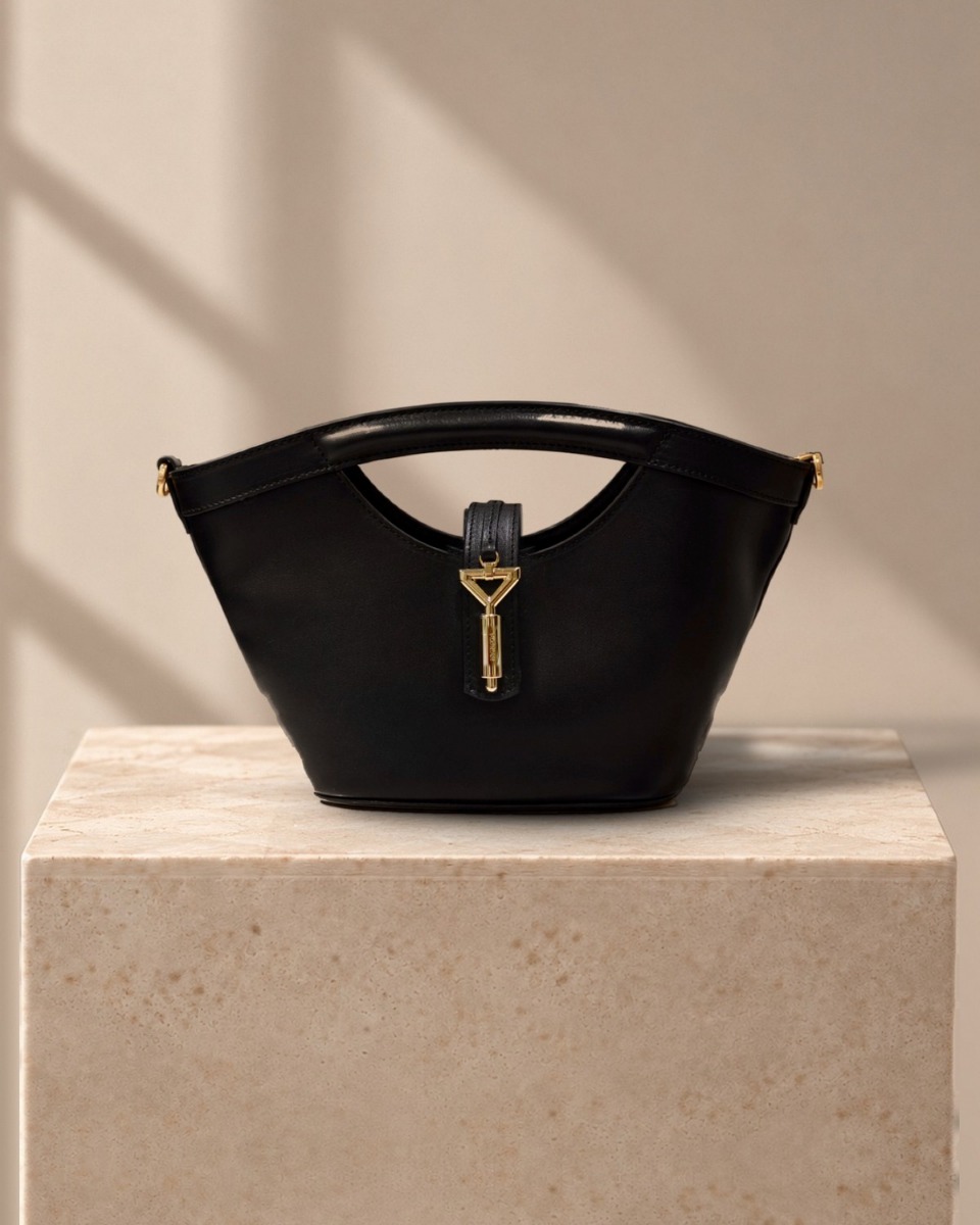 Heidi Tote in black calf leather with gold triangle clasp, on a travertine plinth in warm window light