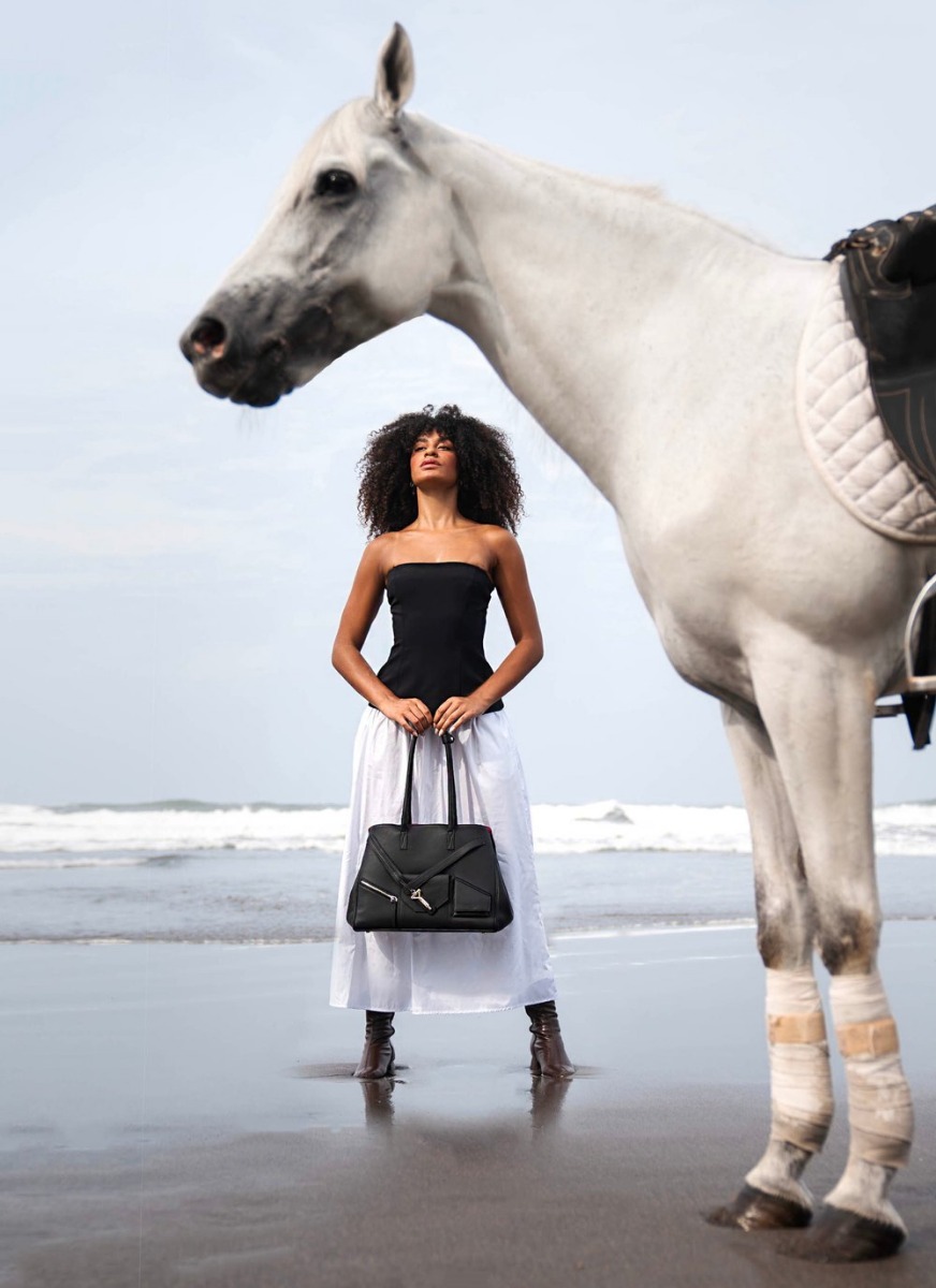 A model in a black strapless top and white skirt on a beach, holding a black Florian Oxford Tote in leather, a white horse in the foreground